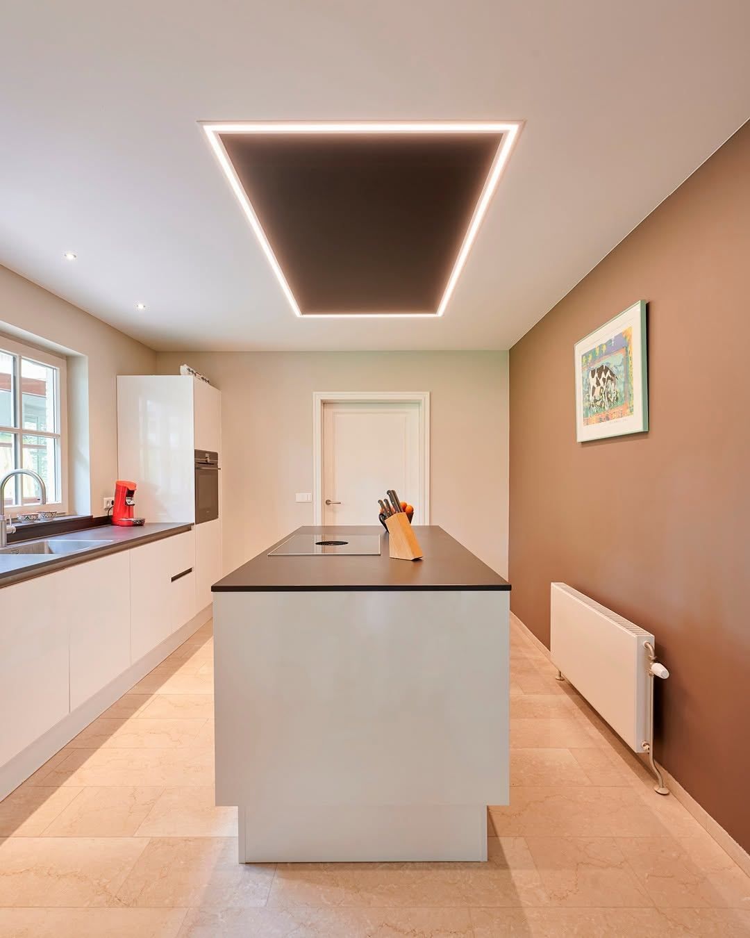 A modern kitchen with an island, a window, and a radiator. The kitchen has a white countertop, a modern ceiling light, and a framed picture on the wall.