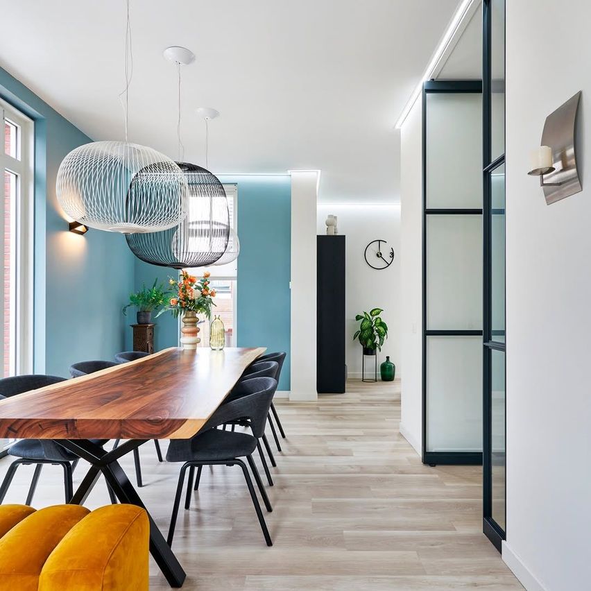 A modern dining room with a long wooden table, black chairs, and blue walls. Three pendant lamps hang from the ceiling. A window and a potted plant are on the right.