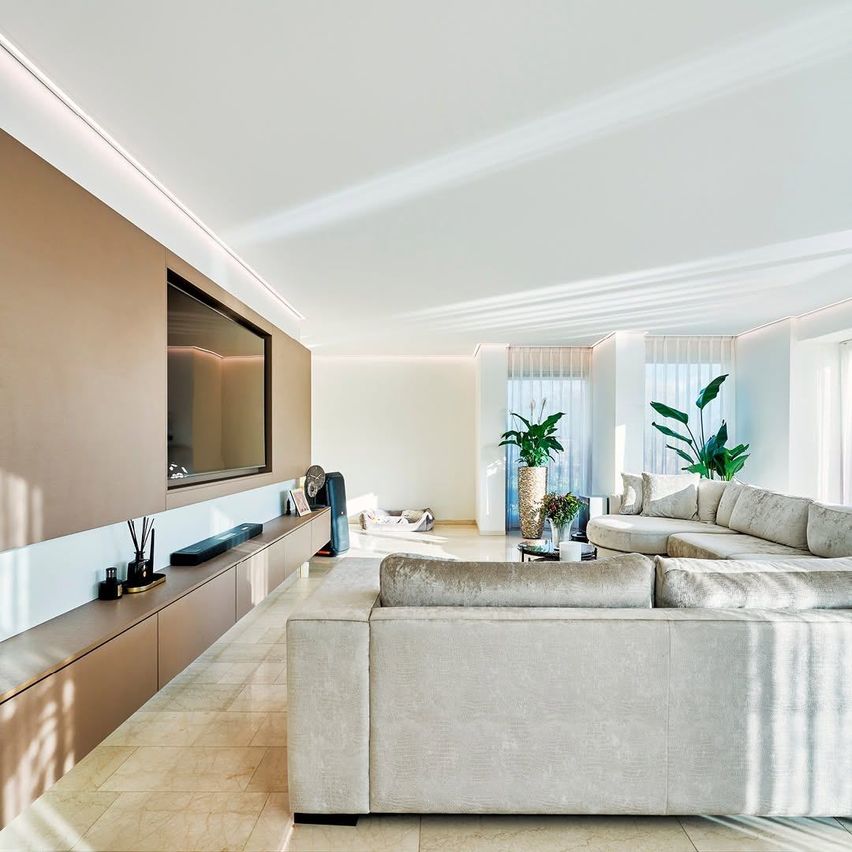A modern living room with a large sectional sofa, a flat-screen TV mounted on the wall, and potted plants. The room has white walls, beige flooring, and an elegant design.