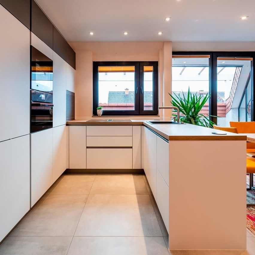 Modern kitchen with white cabinets, beige flooring, and large windows. A plant is on the counter, and a window offers a view of the outside.