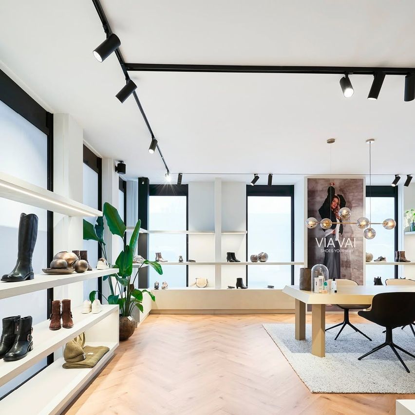 A modern shoe store with white walls and wood floors. Boots, hats, and bags are displayed on shelves. A desk with a black chair sits on a rug in the center.