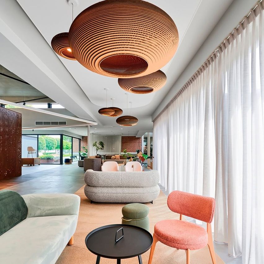A modern lobby area with beige couches, a black table, and wooden chairs, featuring decorative ceiling lamps and white curtains.