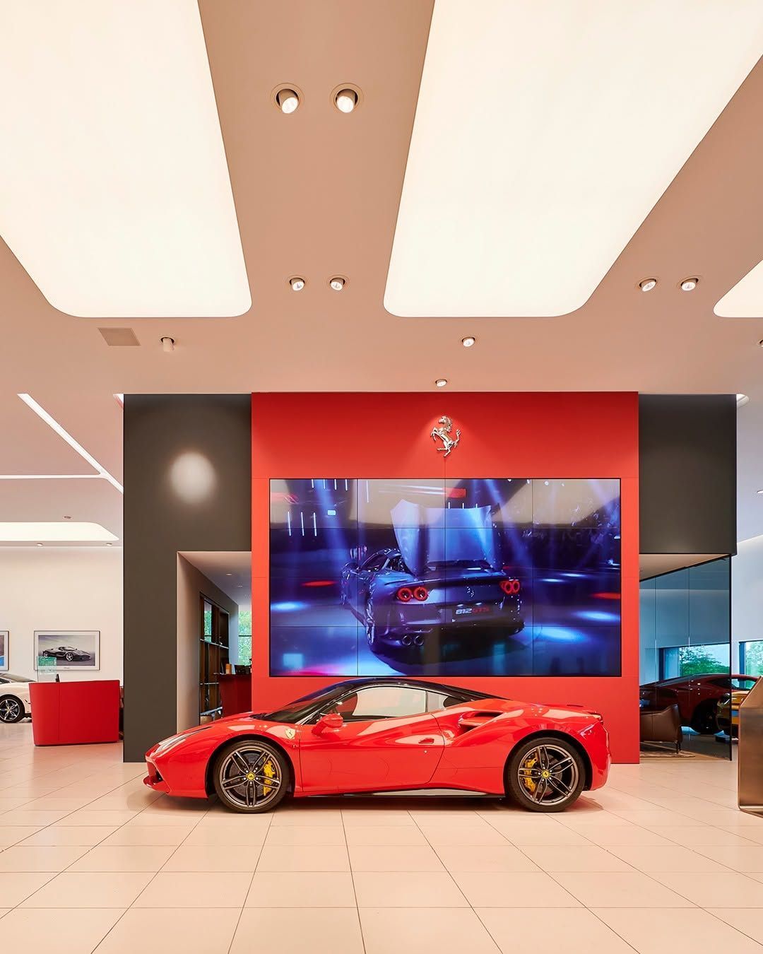 A showroom displays a red Ferrari sports car under a large screen showing a similar car, surrounded by modern furnishings and framed pictures.