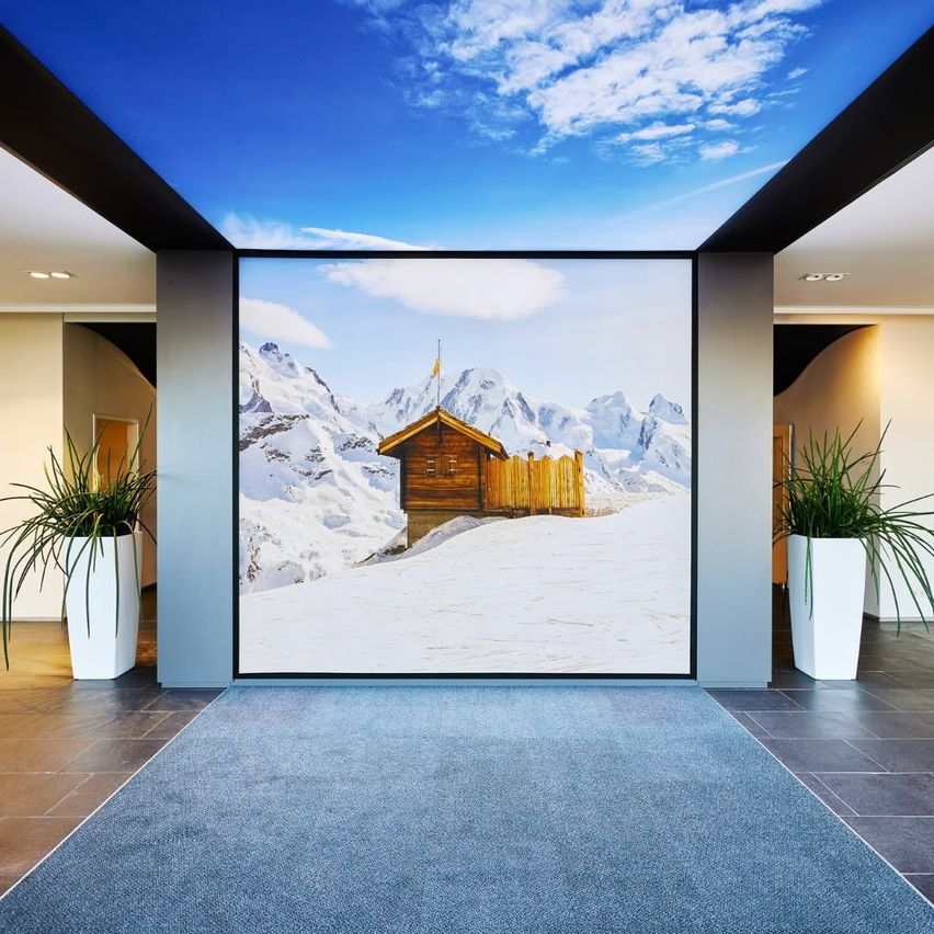 Ein Raum mit einem großen Wandbild einer verschneiten Berglandschaft mit einem kleinen Holzhaus. Zwei weiße Topfpflanzen stehen zu beiden Seiten des Wandbildes.