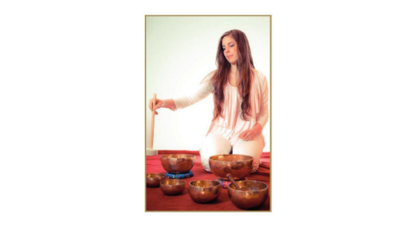 Eine Frau sitzt auf einem roten Teppich und spielt eine Reihe von Bronzegongs mit einem Stock. Die Gongs liegen auf dem Teppich und die Frau schaut nach unten.