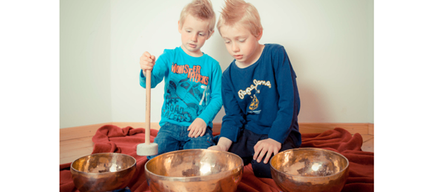 Zwei junge Jungen sitzen auf dem Boden und spielen mit Bronzegongs. Ein Junge hält einen Schläger und klopft sanft auf einen Gong, während der andere Junge ihn ansieht.