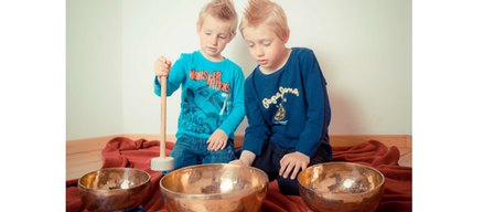 Zwei junge Jungen sitzen auf einem roten Tuch und halten Holzschlägel in der Hand. Sie spielen Kupfergongs. Der Junge auf der linken Seite konzentriert sich darauf, den Gong mit einem Schlägel zu treffen, während der Junge neben ihm aufmerksam zuhört.