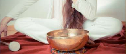 Eine Frau in weißen Kleidern sitzt im Schneidersitz auf einem roten Tuch und blickt auf einen Kupfertopf mit einem kleinen Stock.