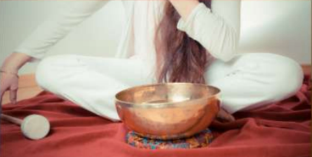 Eine Frau in weißen Kleidern sitzt im Schneidersitz auf einem roten Tuch und blickt auf einen Kupfertopf mit einem kleinen Stock.