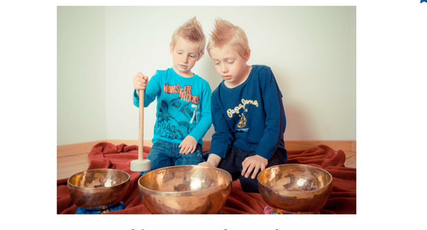 Zwei kleine Jungen sitzen kreuzbeinig auf einer roten Decke, jeder hält einen Holzhammer und ist von Kupfer-Singing-Bowls umgeben. Ein Junge sieht konzentriert aus, während der andere neugierig wirkt. Der Raum hat eine weiße Wand im Hintergrund.