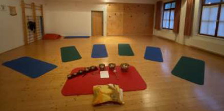 Ein leerer Raum mit Holzböden, Wänden und Decke. Es gibt mehrere Yogamatten und Schüsseln in einem Kreis angeordnet. Ein Kissen ist auf einer roten Matte platziert. Es gibt drei Fenster mit braunen Vorhängen.