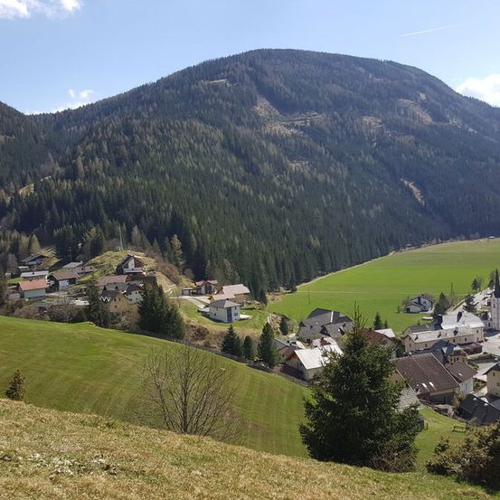 Eine bergige Landschaft mit einem Dorf, das zwischen den Bäumen liegt. Der Himmel ist klar und blau.