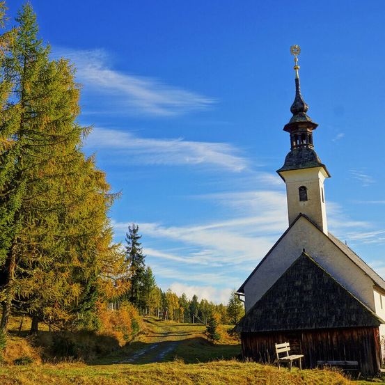 Eine kleine Kirche mit einem Turm und Glockenturm steht auf einem Hügel. Die Kirche ist von Bäumen umgeben, wobei ein hoher Baum im Vordergrund steht. Vor der Kirche steht eine Bank. Der Himmel ist blau mit einigen Wolken.