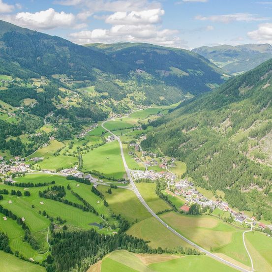 Luftaufnahme eines Tals mit grünen Feldern, einer kurvenreichen Straße und einem kleinen Dorf, eingebettet zwischen den Bergen, unter einem bewölkten Himmel.