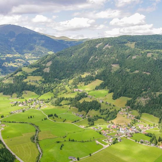 Luftaufnahme eines malerischen Tals mit üppigen grünen Wiesen, einem kleinen Dorf und von Kiefern bewaldeten Bergen unter einem teilweise bewölkten Himmel.