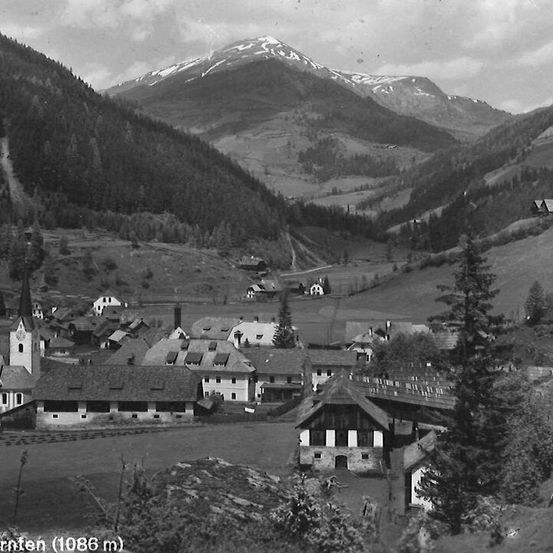 Eine Schwarz-Weiß-Aufnahme eines Bergdorfs mit schneebedeckten Gipfeln, traditionelle Steingebäude und eine Kirche mit einem Turm.