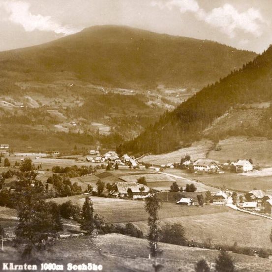 Ein altes Foto zeigt eine bergige Landschaft mit einem Dorf darunter, eingebettet zwischen Bäumen und Feldern. Die Szene wurde in Kärnten in 1000m Höhe über dem Meeresspiegel aufgenommen.