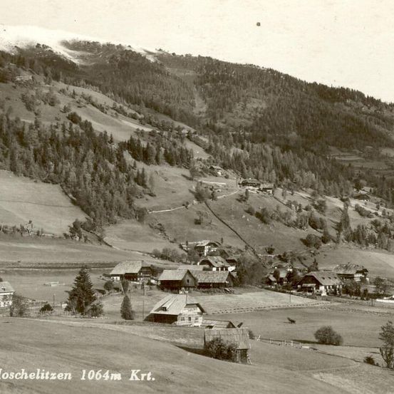 Ein altes Foto eines Dorfes namens Moschelitzen, das auf 1064 Metern über dem Meeresspiegel liegt. Es zeigt mehrere traditionelle Häuser, eingebettet in einem bergigen und bewaldeten Gebiet.