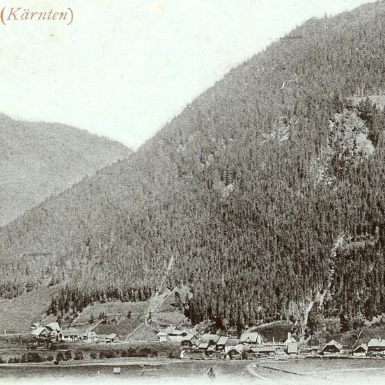 Ein altes Foto zeigt ein Dorf in einem Tal, umgeben von dichten Wäldern und Bergen. Die Region wird als Karnten identifiziert.