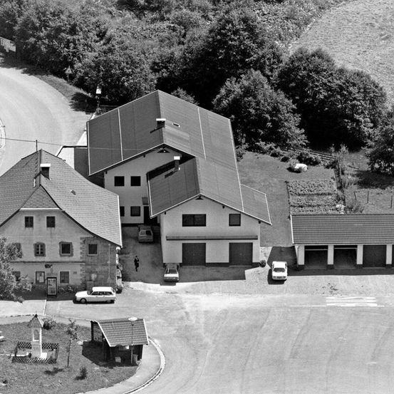 Eine Schwarz-Weiß-Luftaufnahme eines Wohngrundstücks mit mehreren Gebäuden, eines mit grauem Dach. Vor und am Straßenrand sind Autos geparkt.