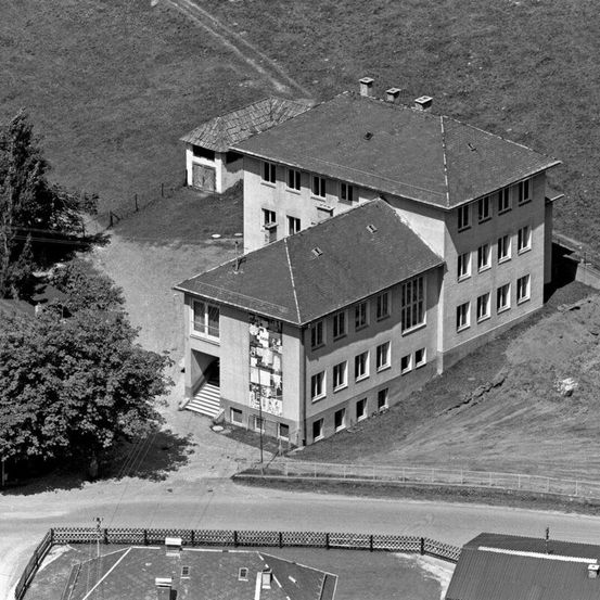 Eine Schwarz-Weiß-Luftaufnahme zeigt ein großes zweistöckiges Gebäude mit vielen Fenstern. Es hat ein flaches Dach und ist von einem eingezäunten Hof umgeben. Bäume und ein Feld sind im Hintergrund.