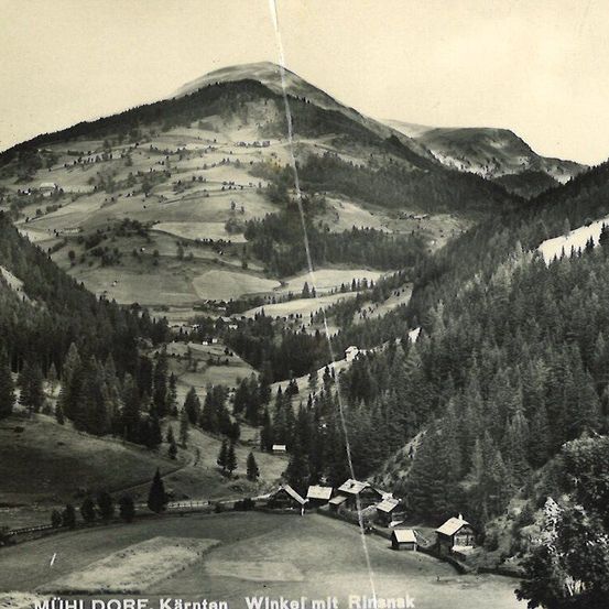 Ein Luftbild einer bergigen Region mit dem Dorf Muhldorf im Vordergrund. Die Landschaft wird von dichten Wäldern und schneebedeckten Hügeln dominiert.