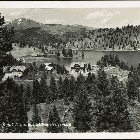 Ein Schwarz-Weiß-Foto zeigt ein friedliches Dorf am See mit einem klaren See, dichten Kiefernwäldern und einem Bergrücken im Hintergrund.