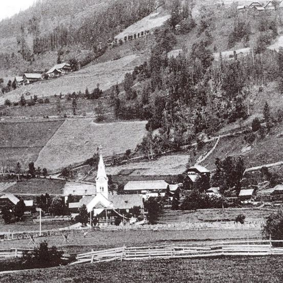 Eine Schwarz-Weiß-Luftaufnahme eines Dorfes mit einer Kirche, umgeben von Hügeln, Bäumen und einem Zaun.