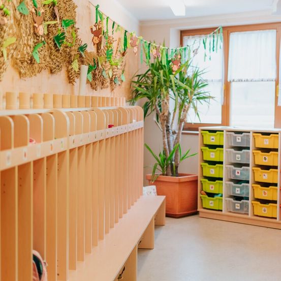 Ein Raum mit Holzregalen und Schuhablagen, einem Topf mit Pflanzen und farbenfrohen Aufbewahrungsboxen. Die Wände sind mit Papiertieren und Grün dekoriert.