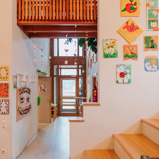 Ein Korridor mit weißen Wänden führt zu einer Tür. Mehrere bunte Kinderbilder schmücken die Wände. Oben befindet sich ein Holzbalkon.
