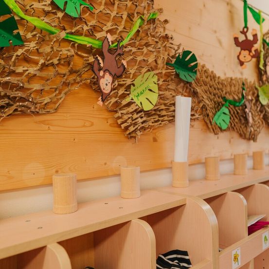 Ein holzverkleideter Klassenraum mit Dschungel-Thema, der eine Wand mit Papierausschnitten von Blättern und Affen zeigt. Es gibt ein Regal mit mehreren hölzernen Ablagen zur Aufbewahrung von Gegenständen.