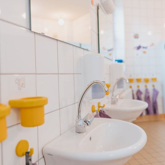 Ein Badezimmer mit weißen Fliesen, zwei Waschbecken, gelben Haltern und lila Handtüchern an der Wand.