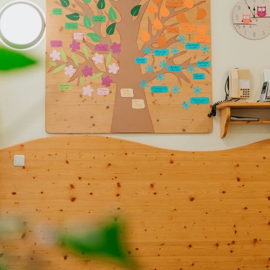 Eine Holzwand mit einem Baumdesign, verziert mit bunten Blättern und Notizen. Darüber eine runde Uhr mit einer Eulenfigur. Rechts nebenan ein Holzregal mit einem Telefon.