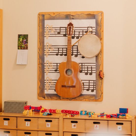 Ein Raum mit einer Wand, die eine gerahmte Gitarre und Musiknoten zeigt, begleitet von einem Trommel und anderen Instrumenten. Hölzerne Schubladen darunter sind mit bunten Spielzeugen gefüllt.
