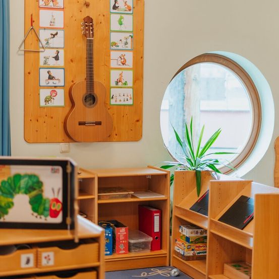 Ein Klassenzimmer mit Holzmöbeln, einer Gitarre und einem runden Fenster. Es gibt eine Pflanze, Bücher und Spielzeug auf Regalen.