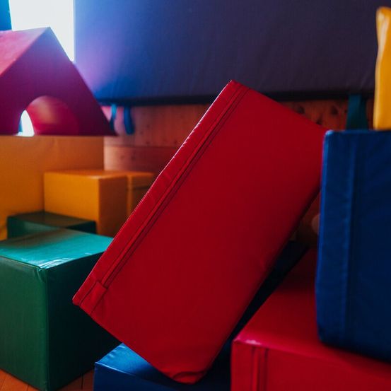 Ein rotes Kissen liegt auf einem blauen und roten Kissen, dahinter sind weitere farbige Kissen und eine hausähnliche Struktur.