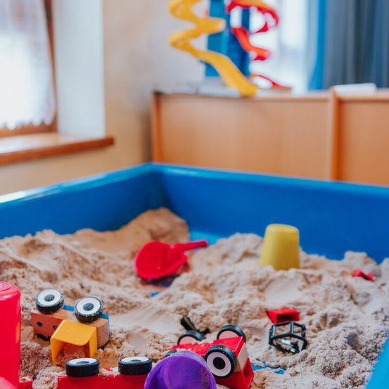 Ein Sandkasten voller Spielzeug, darunter Autos, eine Schaufel und ein Eimer, mit einer bunten Spirale und einem Fenster mit weißen Vorhängen im Hintergrund.