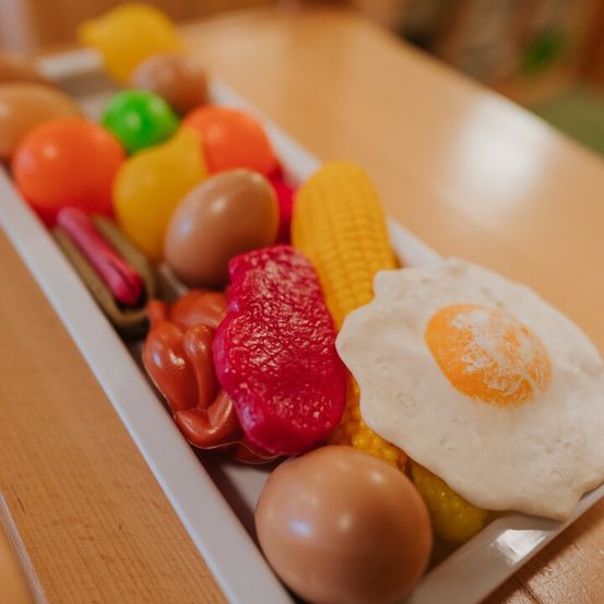 Ein rechteckiger Teller gefüllt mit verschiedenen farbigen lebensmittelähnlichen Objekten, darunter Eier, Mais, Paprika und ein Spiegelei. Der Teller steht auf einem Holztisch.