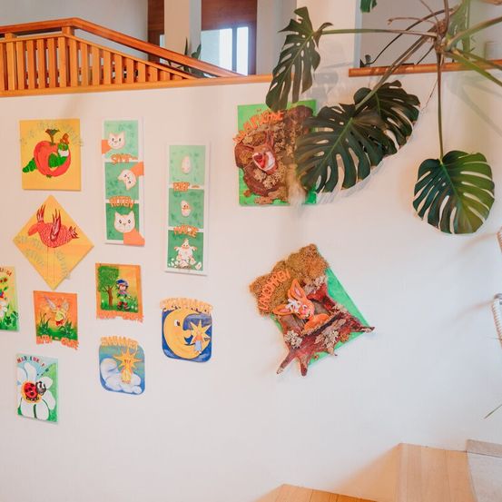 An einer weißen Wand sind verschiedene bunte Gemälde und Illustrationen zu sehen, darunter Kürbisse, Katzen, Eier und eine Sonne. Ein großer grüner Pflanzen hängt von der Wandspitze.