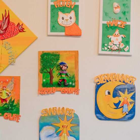 Eine Wand mit verschiedenen Gemälden und Illustrationen. Eines hat einen roten Vogel, ein anderes eine Katze und ein lächelndes Gesicht. Es gibt auch Zeichnungen von einer Fee, einem Jungen und einem Mond mit einem Stern.