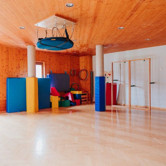 Ein Innenbereich mit Holzwänden und -boden. Verschiedene farbenfrohe Übungsgeräte sind im Raum angeordnet, einschließlich einer blauen Schaukel, die von der Decke hängt.
