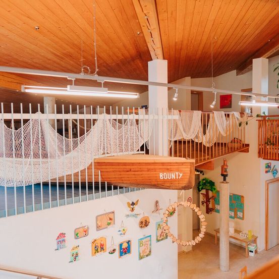 Ein Indoor-Spielbereich mit einem hölzernen Boot namens 'BOUNTY', das darüber hängt. Der Bereich umfasst ein Hängemattenbett, einen Balkon und verschiedene Dekorationen. Lampen sind an der Decke montiert.
