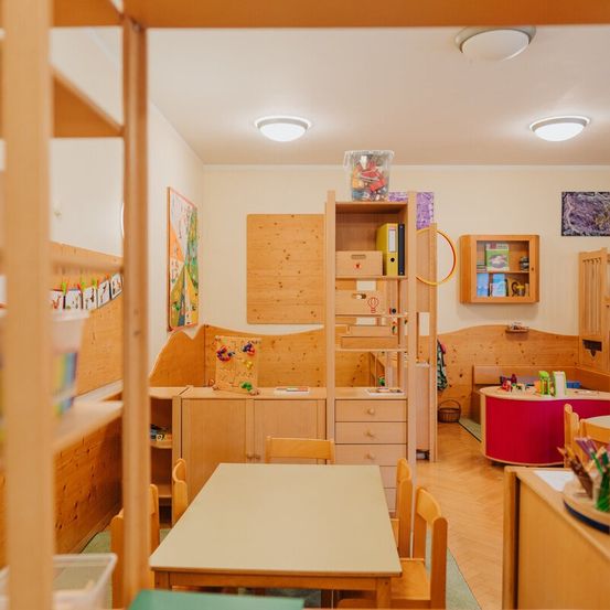 Ein Kinderzimmer mit Holzmöbeln und Spielzeug. Ein Tisch, Stühle und ein Holzregal mit Spielzeug. Ein Bilderrahmen und eine Aufbewahrungsbox hängen an der Wand.