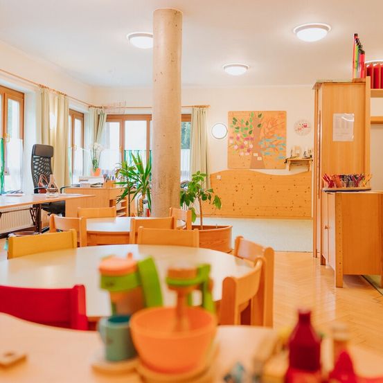 Ein hell beleuchteter Essbereich mit Holztischen und Stühlen. Der Raum hat einen Holzboden und weiße Wände. Es gibt eine zentrale Säule und ein Kunstwerk an der Wand.