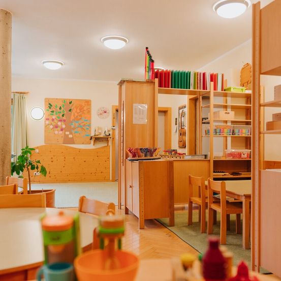 Ein Klassenzimmer mit Holzmöbeln, einem Schreibtisch, Stühlen und einem Regal mit farbenfrohen Ordnern. Ein kreisförmiges Baumbild an der Wand und eine Pflanze in einem Topf. Der Raum hat eine warme Atmosphäre.