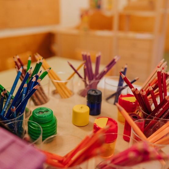 Ein Klassenzimmertisch ist mit verschiedenen farbigen Stiften, Malstiften und Markern gefüllt. Es gibt auch Gläser für Anspitzer und Bleistifte sowie einige Behälter mit Deckeln. Der Raum hat Holzregale und Stühle im Hintergrund.