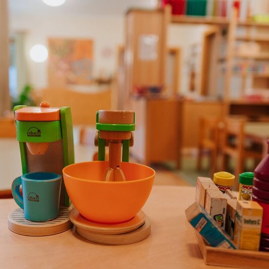 Auf einem hölzernen Tisch ist ein buntes Spielzeug-Küchenset ausgestellt. Das Set enthält eine blaue Tasse, eine orangefarbene Rührschüssel und einen grünen und orangefarbenen Spielzeug-Mixer. Verschiedene Gewürzflaschen sind an der Seite.