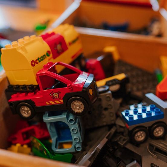 Eine Schachtel mit bunten Spielzeugfahrzeugen, darunter ein rotes Auto mit dem Feuerwehrmann-Emblem, steht auf anderen Spielzeugen. Ein gelber Tankwagen mit 'Octane' steht im Hintergrund.