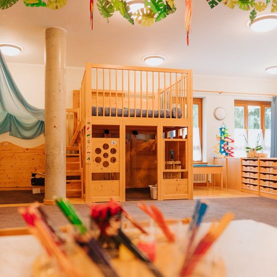 Ein Innenbereich für Kinder mit einem Holzbett und einer Treppe. Der Raum hat Holzschränke, Regale und einen Schreibtisch. Spielzeug und Gegenstände sind auf dem Boden verstreut. Es gibt eine weiße Wand mit einer Uhr und blauen Vorhängen.