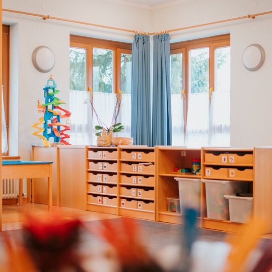 Ein Klassenzimmer mit weißen Wänden, Holzmöbeln und blauen Vorhängen. Reihen von Schubladen und Aufbewahrungsboxen enthalten Spielzeug und Lernmaterialien. Ein Spiralspielzeug steht auf einem Regal.